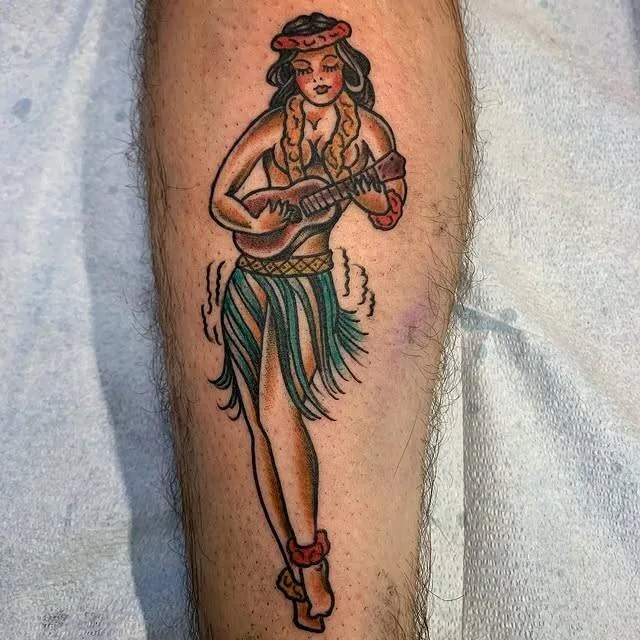 American Traditional hula-girl pin-up tattoo on a calf — woman in lei, grass skirt, and red heels strumming a ukulele — by Forrest Goodrich at Ohana Tattoo, Waikiki, Hawaii.