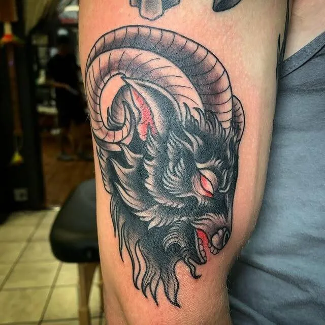 Black and grey horned ram tattoo with glowing red eye and open mouth on an upper arm, by Forrest Goodrich at Ohana Tattoo, Waikiki, Hawaii.