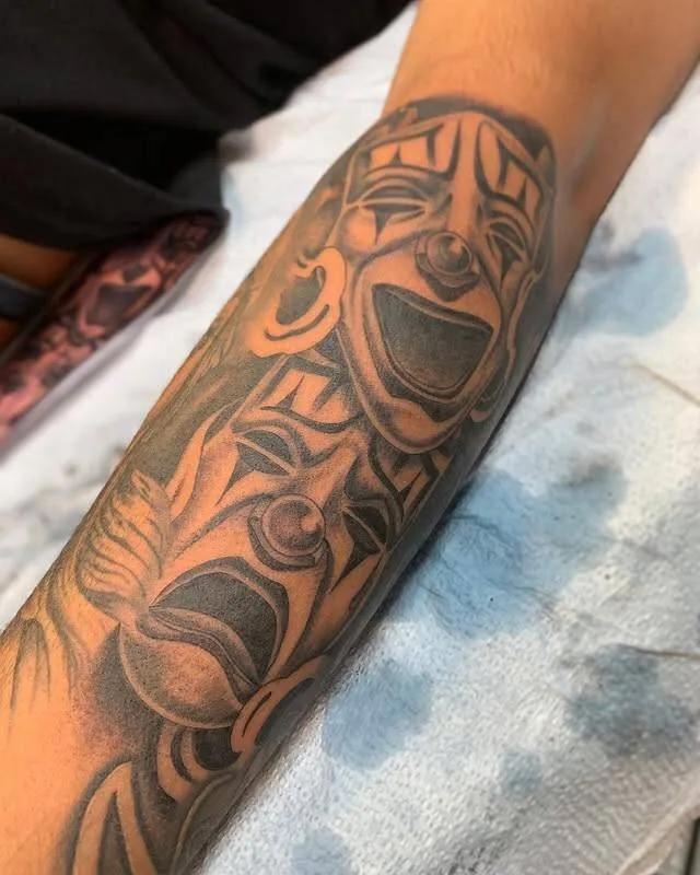Forearm sleeve tattoo blending a laughing black-and-grey jester or theater mask with bold Polynesian tribal framing, by Forrest Goodrich at Ohana Tattoo, Waikiki, Hawaii.