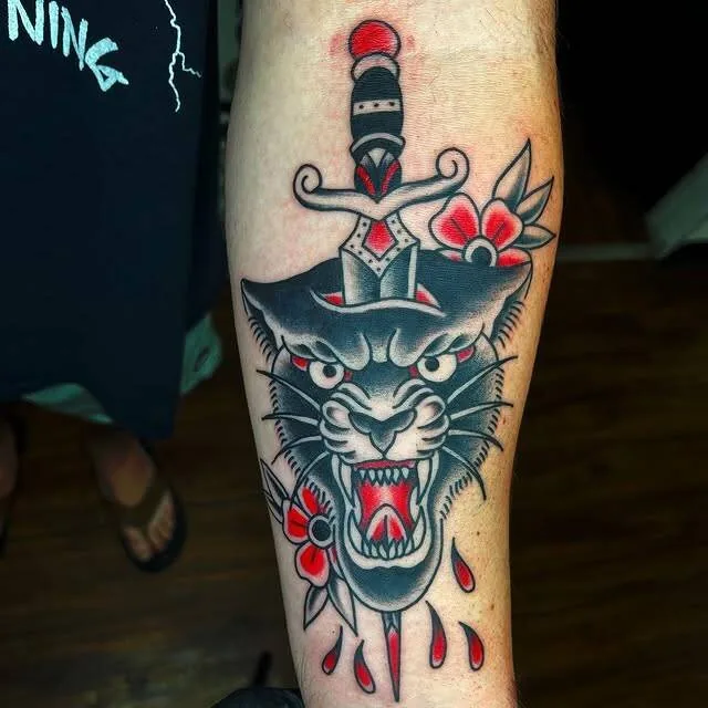 American Traditional panther head tattoo on a forearm, pierced from above by an ornate dagger with red cherry blossoms and dripping blood drops, by Forrest Goodrich at Ohana Tattoo, Waikiki, Hawaii.