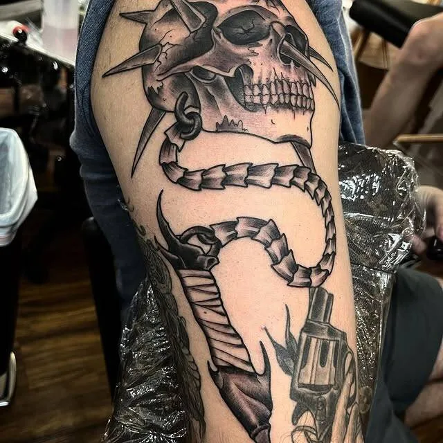 Black and grey half-sleeve tattoo of a spiked skull with a long trailing spine and a revolver underneath, by Forrest Goodrich at Ohana Tattoo, Waikiki, Hawaii.
