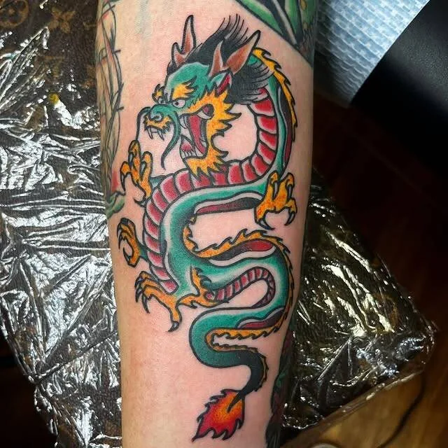 Traditional Asian dragon tattoo on a forearm in jade green and saturated orange, claws extended and tail flame trailing, by Forrest Goodrich at Ohana Tattoo, Waikiki, Hawaii.