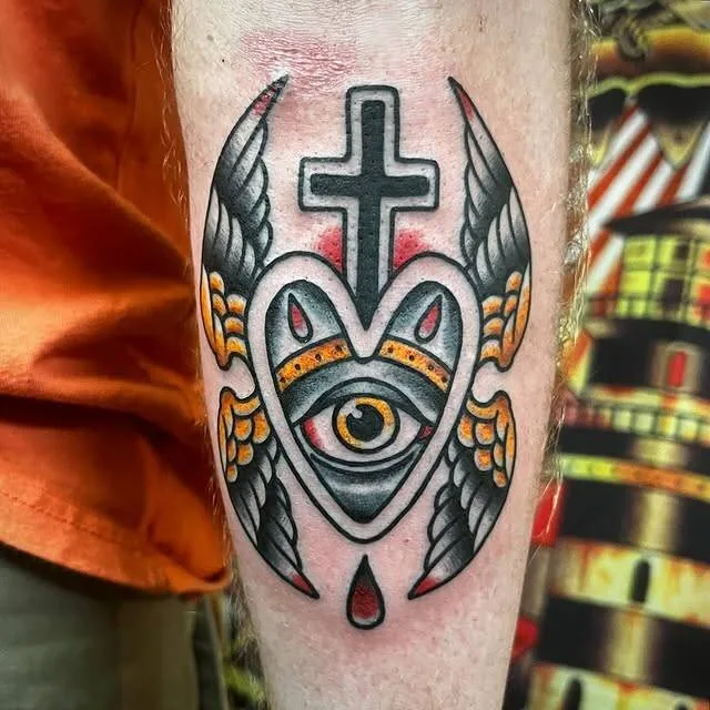 American Traditional sacred-heart tattoo with the all-seeing eye in the center, flanked by wings and capped with a cross, on a calf, by Forrest Goodrich at Ohana Tattoo, Waikiki, Hawaii.