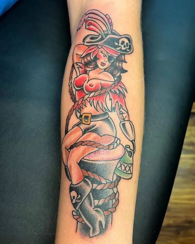 American Traditional pirate pin-up tattoo on a forearm — woman in eye patch, tricorn hat, fur boots holding a rum bottle and coiled rope — by Forrest Goodrich at Ohana Tattoo, Waikiki, Hawaii.